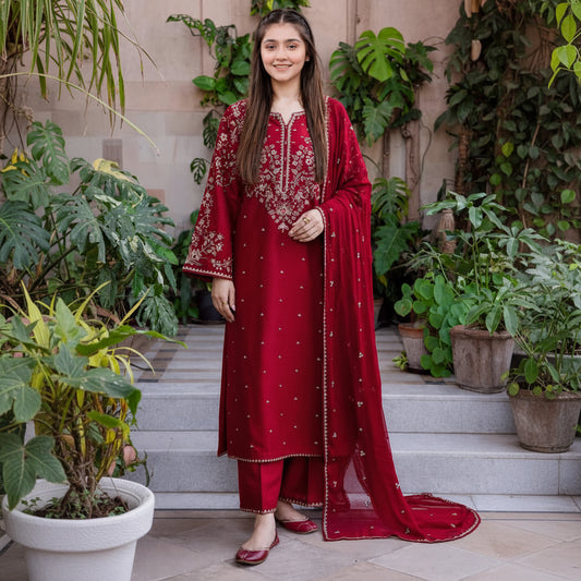 A woman modeling a deep red Indian ethnic suit with intricate gold embroidery, matching trousers, and a sheer dupatta, standing on stone steps surrounded by green plants.