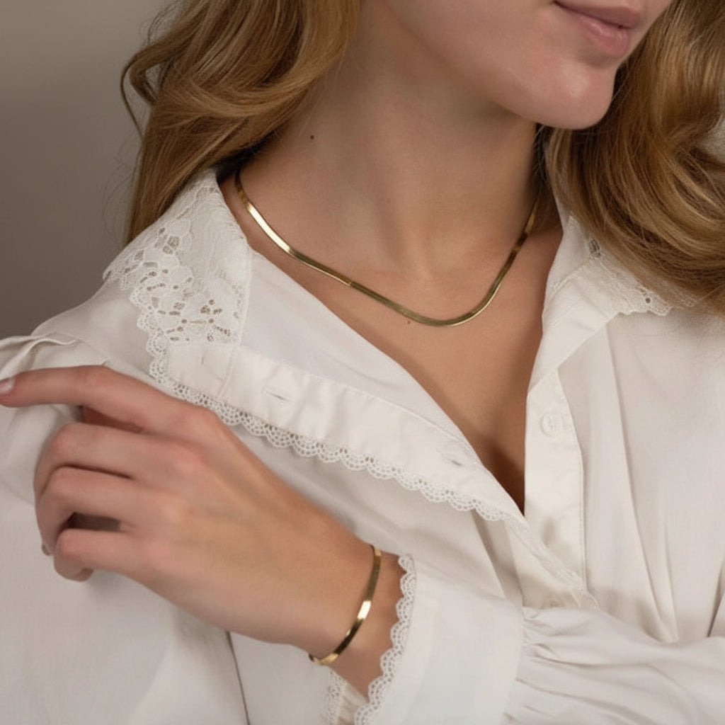 A close-up lifestyle shot of a woman wearing a delicate gold snake chain necklace and a matching gold bangle bracelet, paired with a white lace-trimmed blouse.