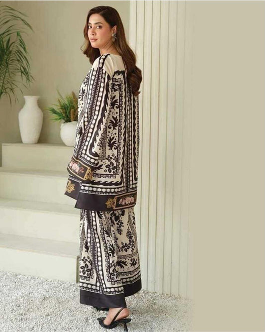 A woman models a three-piece printed ethnic suit in a neutral indoor setting. The suit features a cream base with black floral patterns and brown borders, paired with a matching long jacket and wide-leg trousers.