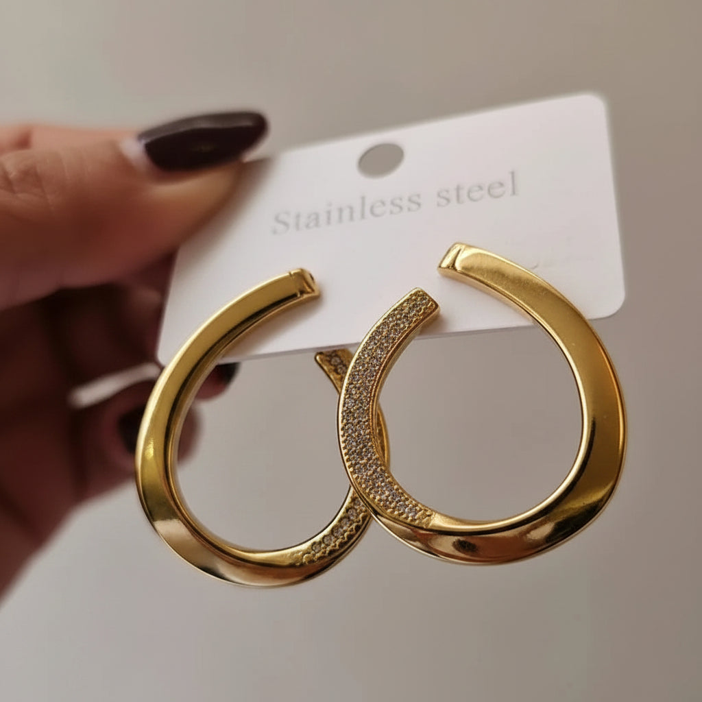 A close-up of a hand holding a pair of gold-tone open hoop earrings with a curved row of small clear stones, attached to a white card labeled 'Stainless steel.'.