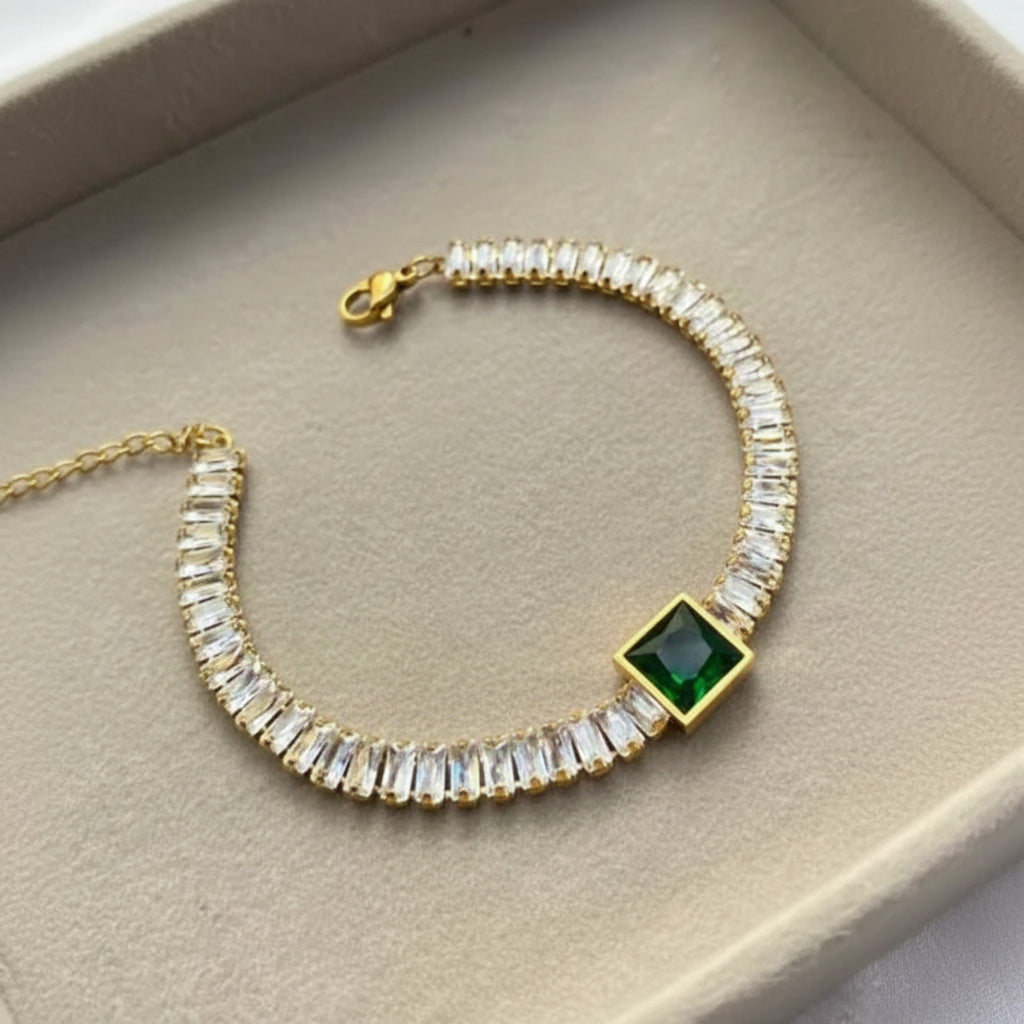 A gold-plated tennis bracelet featuring clear rectangular crystals and a central square green gemstone, displayed inside a beige jewelry box.