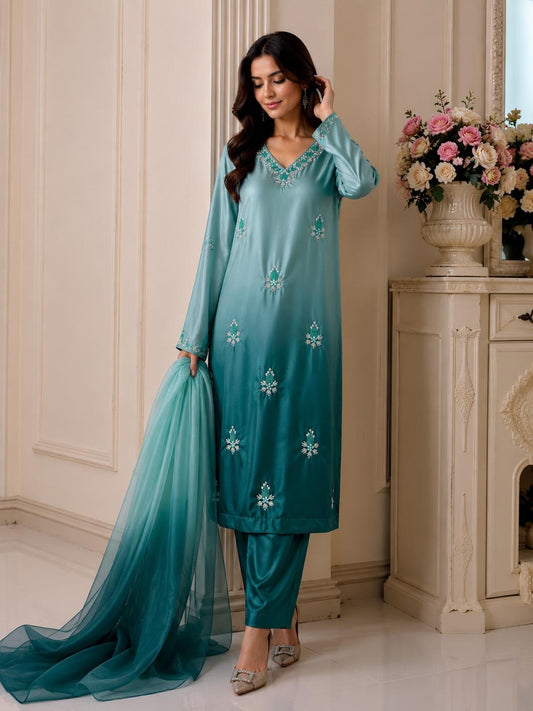 A woman models a teal blue-green gradient Indian ethnic suit with matching pants and a long sheer dupatta, standing in a classic cream-colored room next to a floral console table.
