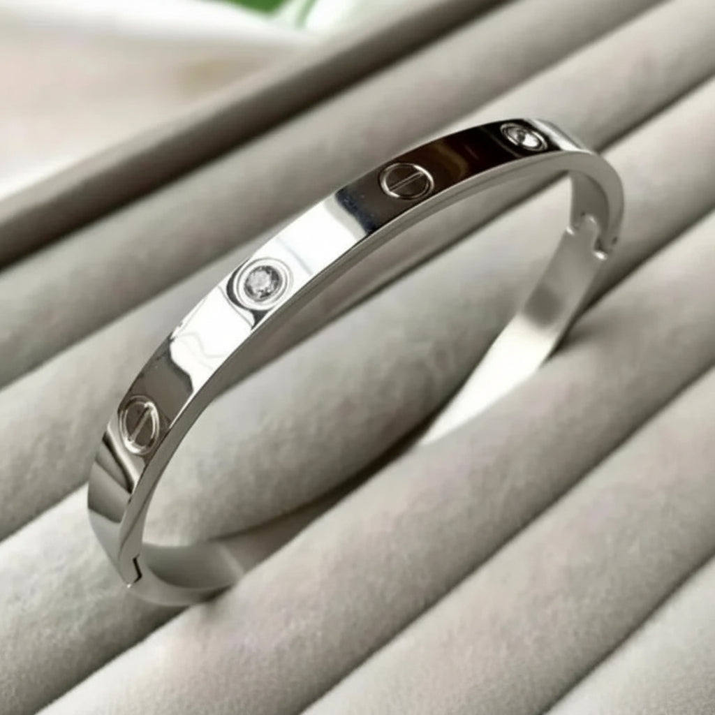 A silver-toned bangle bracelet with three screw-head details and small clear stones, displayed on a light gray velvet jewelry organizer.