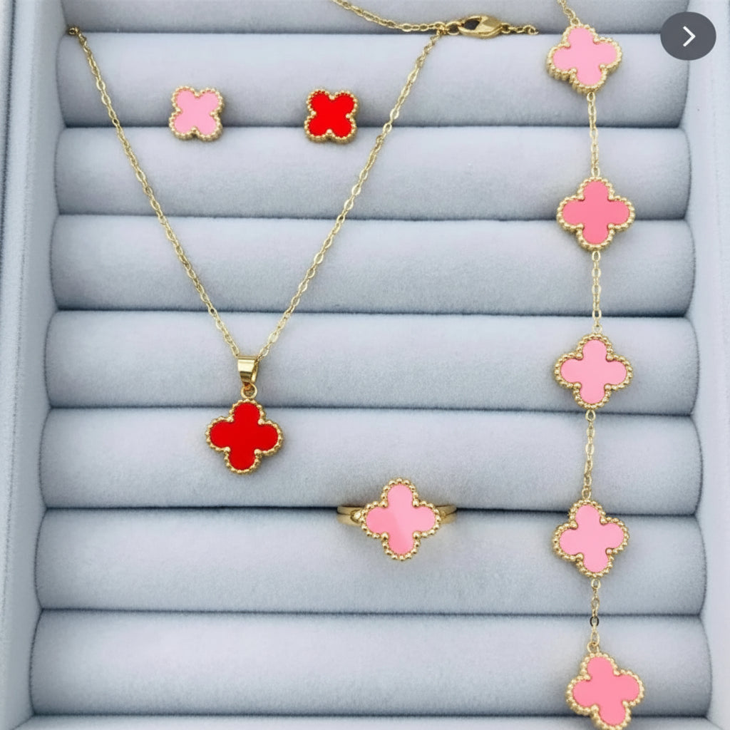 Gold jewelry set with red clover-shaped enamel details, including a necklace, ring, earrings, and bracelet, displayed on a light blue-gray padded jewelry box.