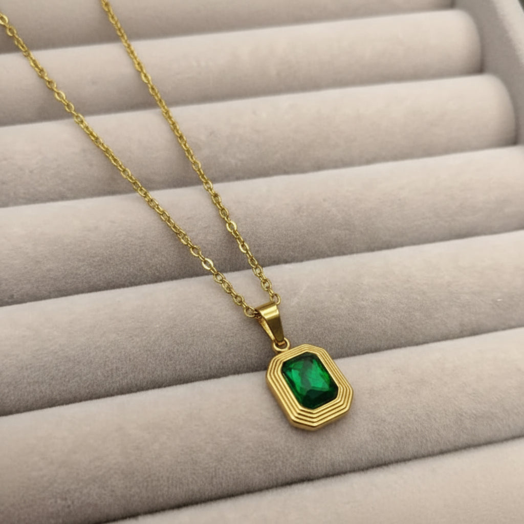 A gold chain necklace with a gold geometric pendant featuring a green rectangular gemstone, displayed on a gray padded jewelry organizer.