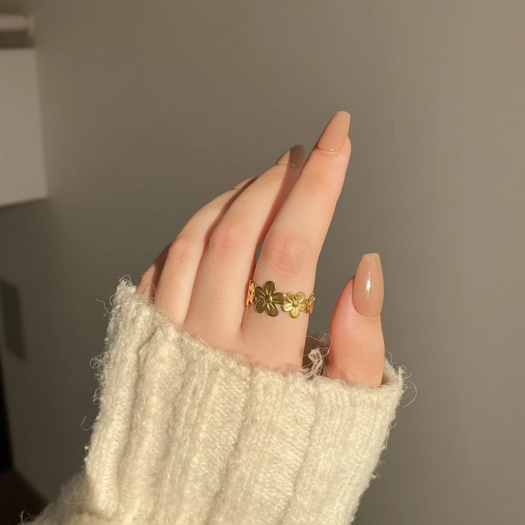 A close-up of a hand wearing a gold ring with three small flower motifs, styled with a cream-colored knit sweater sleeve.