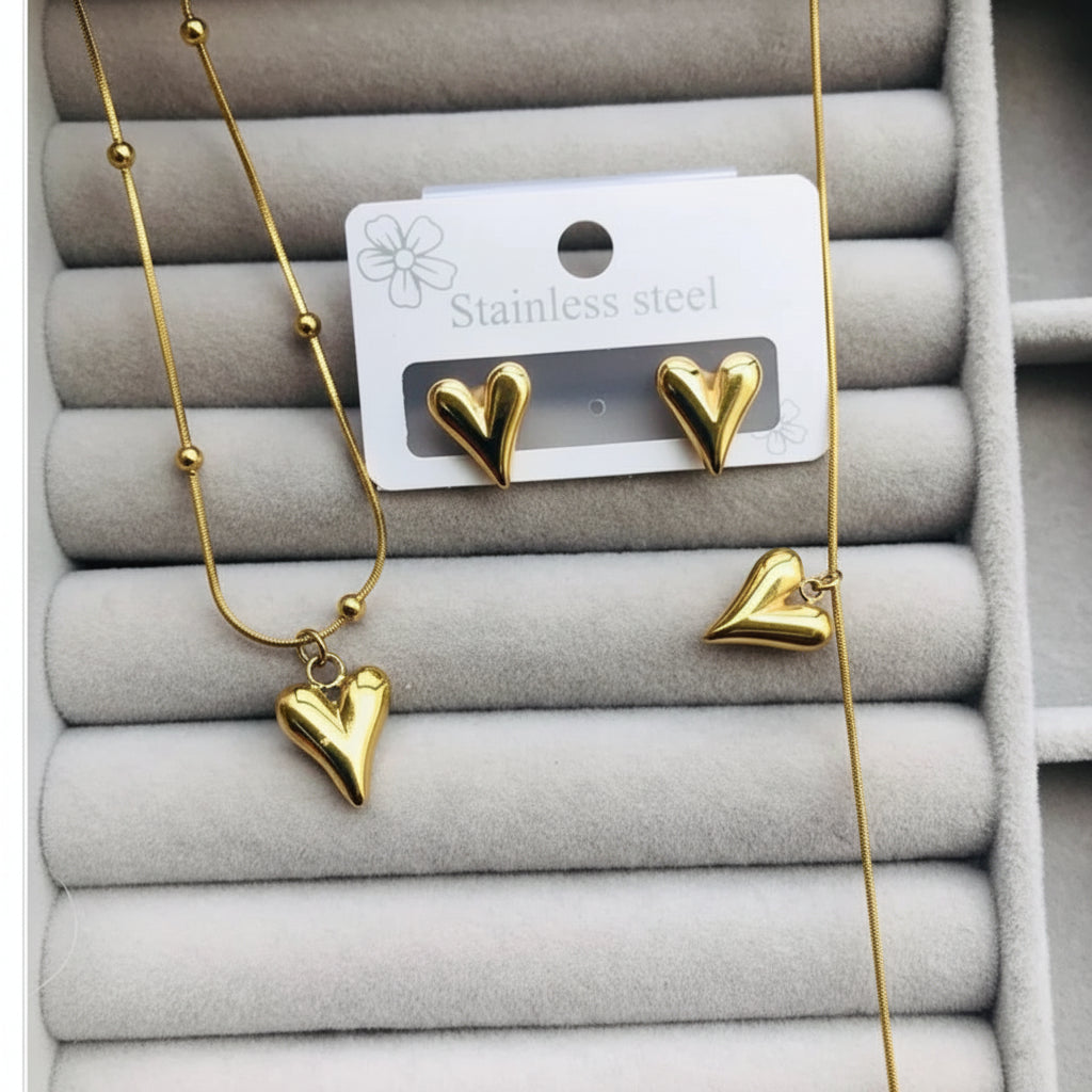 Gold heart jewelry set including a necklace, stud earrings, and a heart-shaped pendant, displayed on a gray jewelry tray with a white card labeled 'Stainless steel'.