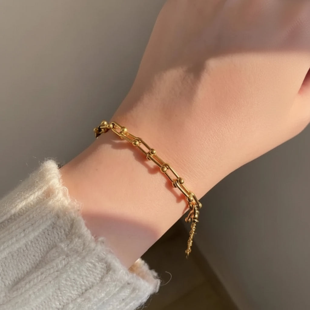A gold chain bracelet with a small dangling charm, shown worn on a person's wrist against a neutral indoor background.
