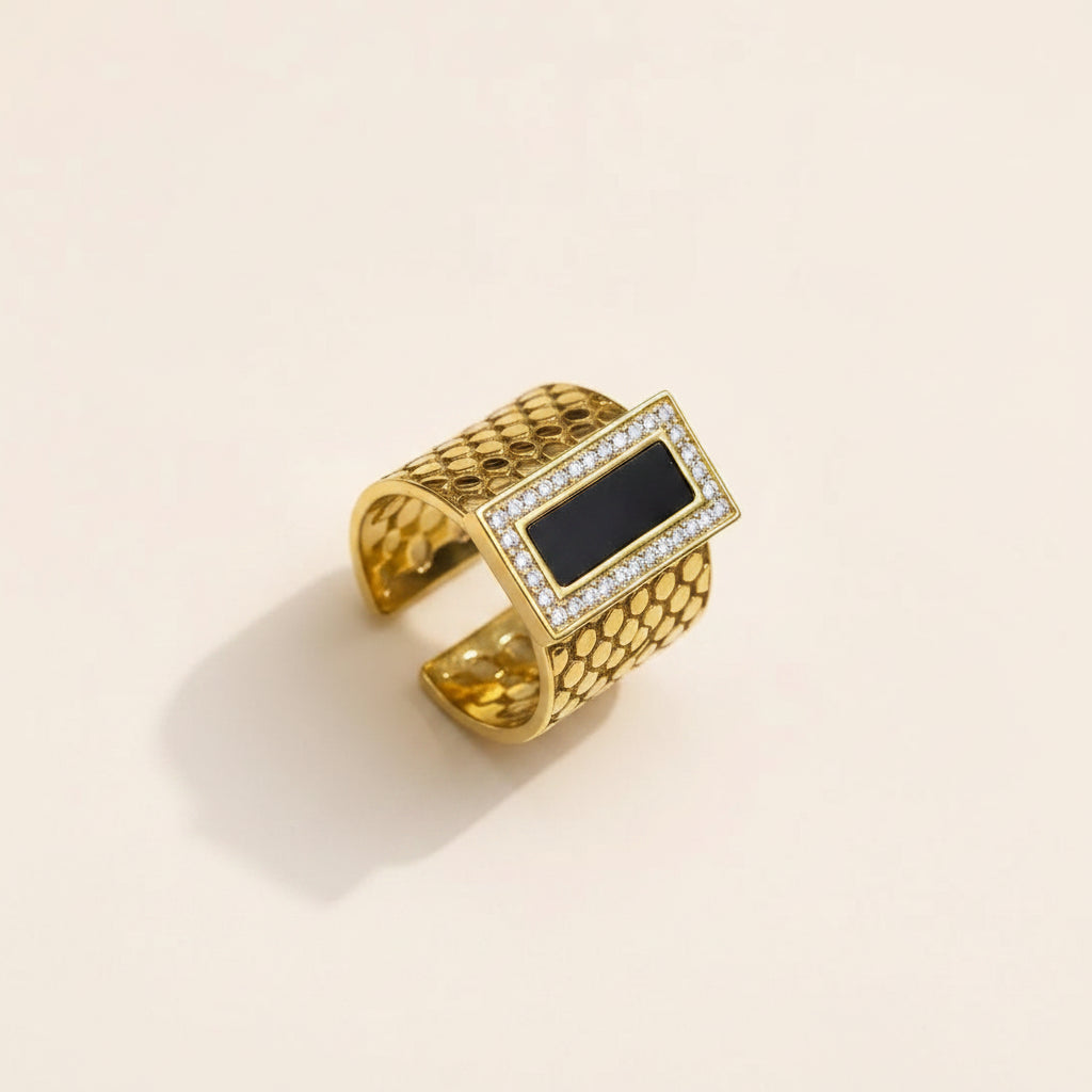 A gold-toned open cuff ring featuring a rectangular black inlay bordered by small clear stones, photographed on a light beige background.