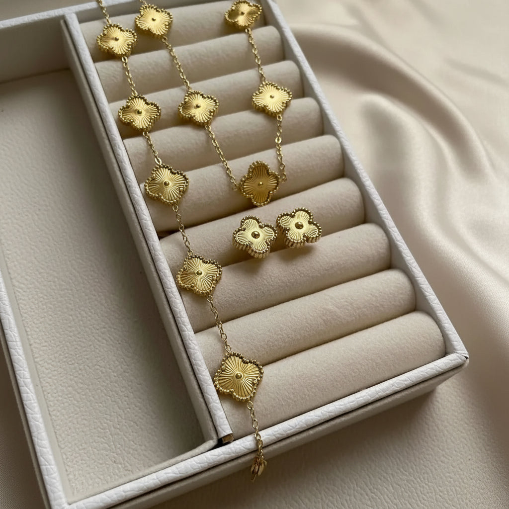 Gold Alhambra-style flower necklace and matching stud earrings displayed on a cream jewelry box. The necklace features multiple textured gold flower motifs connected by a delicate chain, and the earrings are matching flower-shaped studs with small central gem accents.