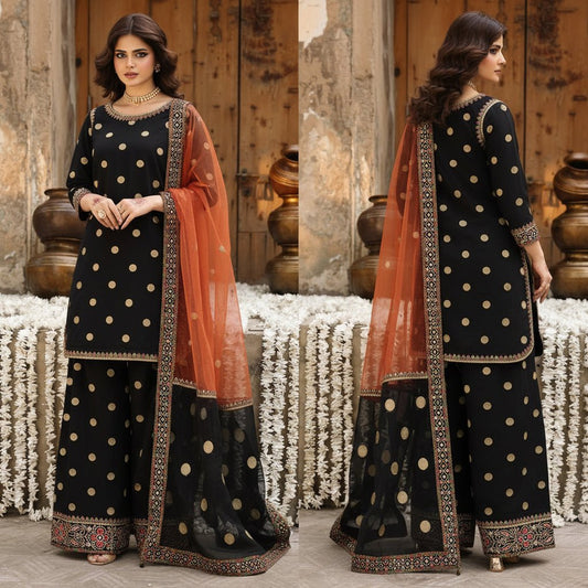 Woman in a traditional black and gold outfit with an orange dupatta standing against a rustic wooden background.