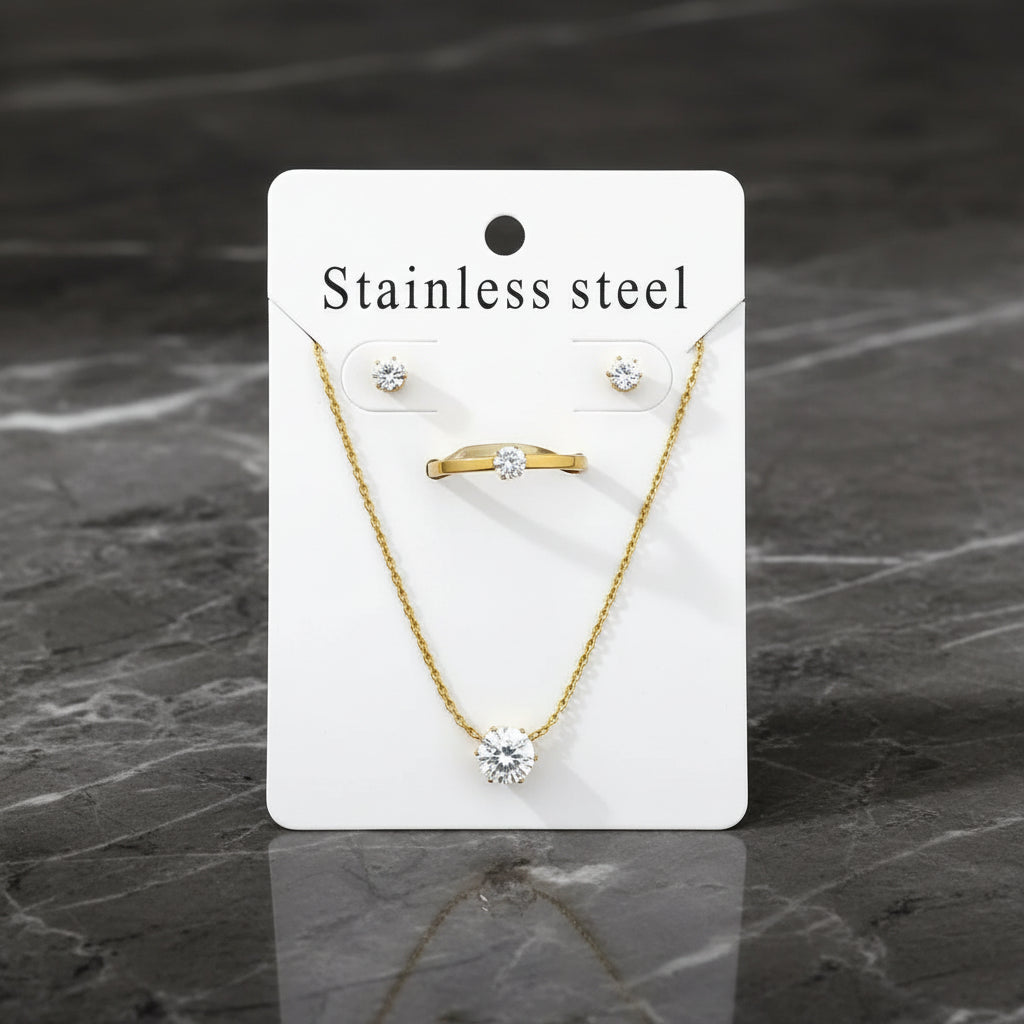 A jewelry set displayed on a white backing card labeled 'Stainless steel,' including a gold-tone ring with a clear stone, a gold-tone necklace with a round clear stone pendant, and matching stud earrings. The card is photographed on soft white fabric.