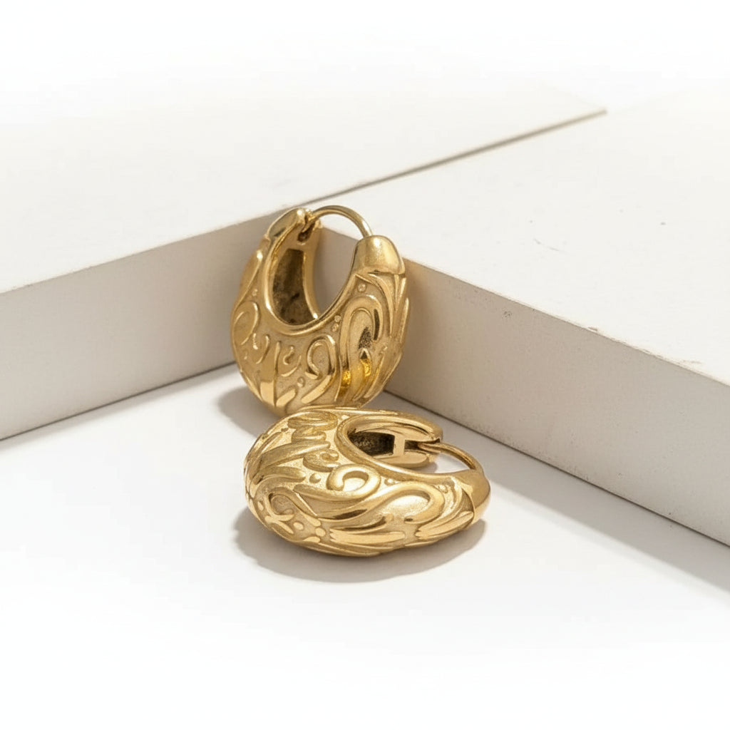 A pair of gold-toned, heavily textured and ornate hoop earrings displayed on a white geometric riser against a dark background.