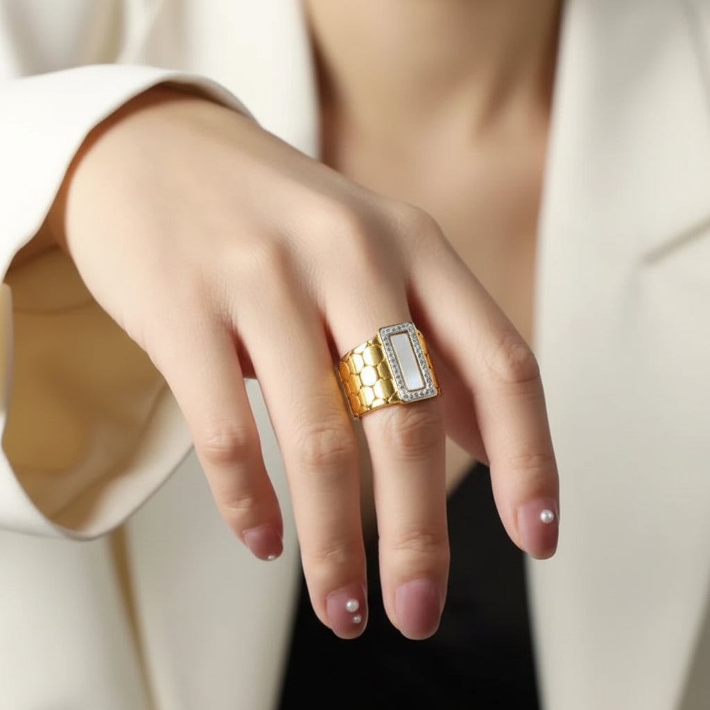 A close-up of a person's hand wearing a gold and silver ring with a rectangular white inlay, styled with a white blazer.