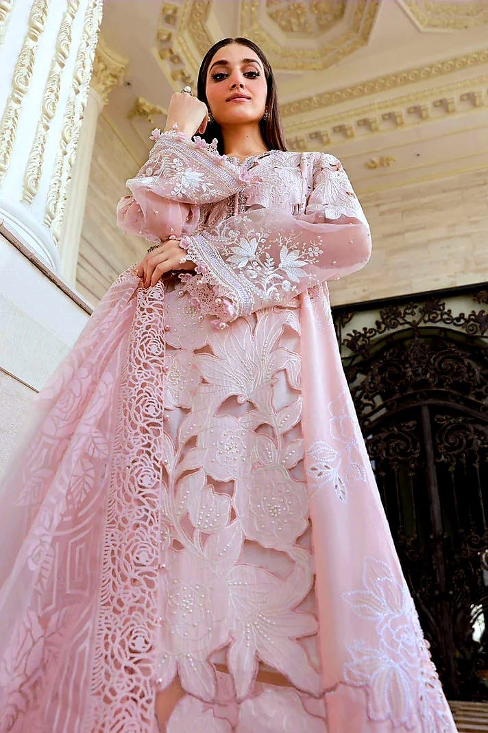 A woman wearing a voluminous light pink traditional dress with intricate floral embroidery and sheer lace panels, posed in front of an ornate beige building interior.