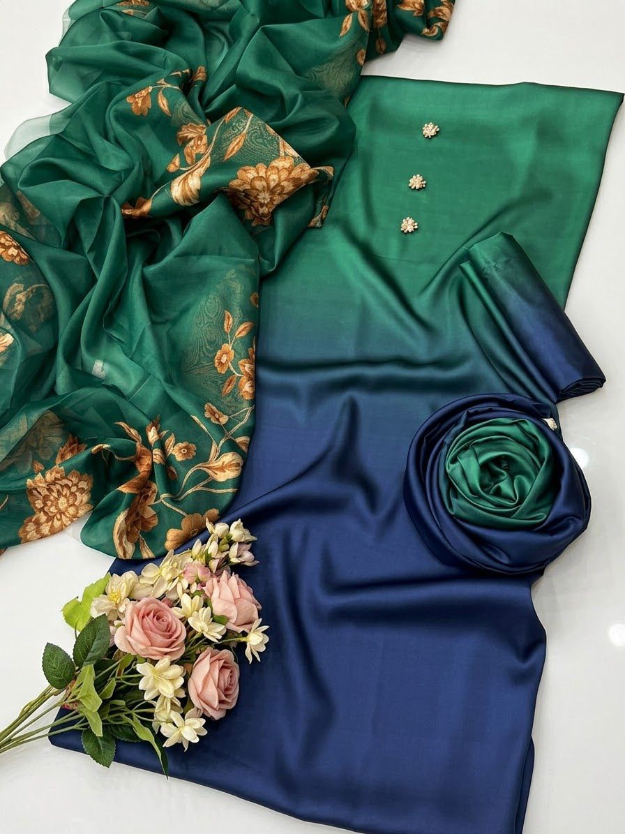 A folded and draped green satin fabric set with a gradient transitioning to deep blue, accompanied by a sheer green dupatta with gold floral patterns and a small bouquet of pink and white roses on a white surface.