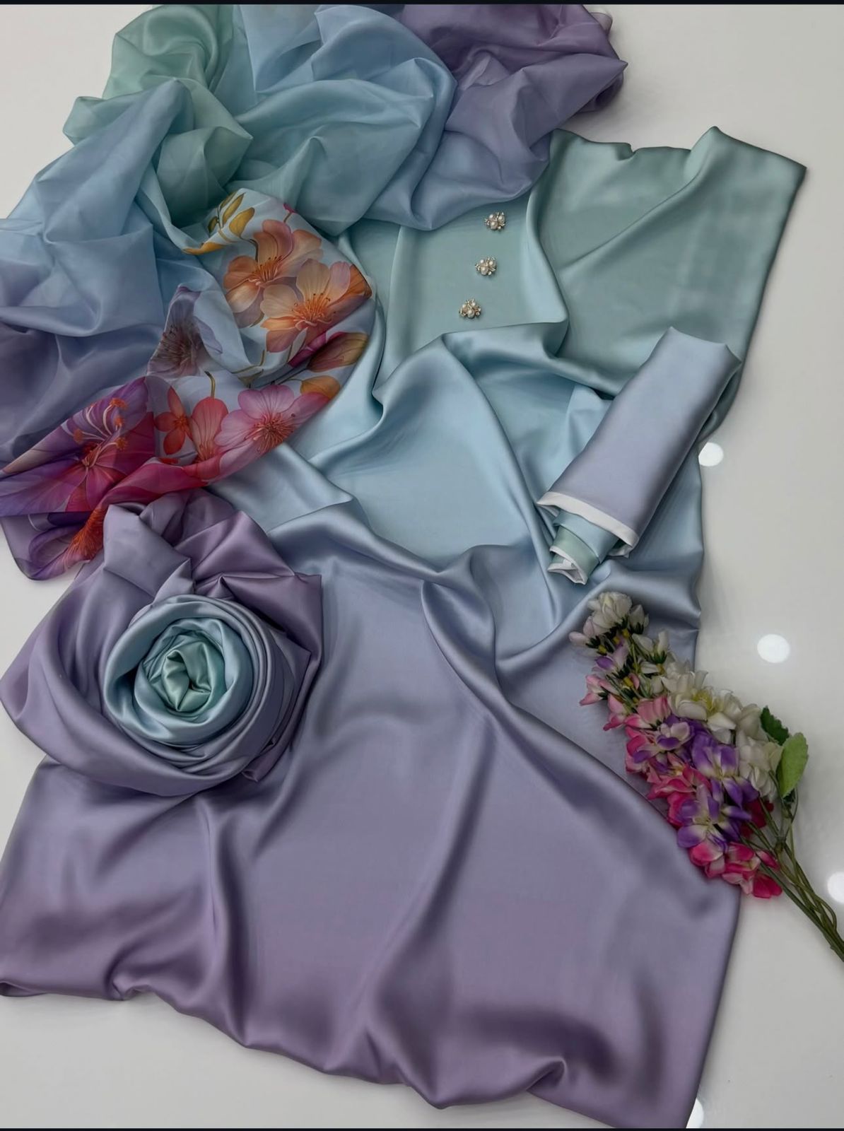 A styled flat-lay of light blue and lavender satin fabric, including a draped dress or garment, a matching scarf with a floral print, a rolled fabric piece, a fabric rose, and a small bouquet of flowers on a white surface.