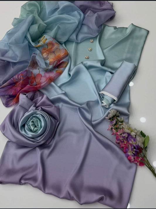 A styled flat-lay of light blue and lavender satin fabric, including a draped dress or garment, a matching scarf with a floral print, a rolled fabric piece, a fabric rose, and a small bouquet of flowers on a white surface.