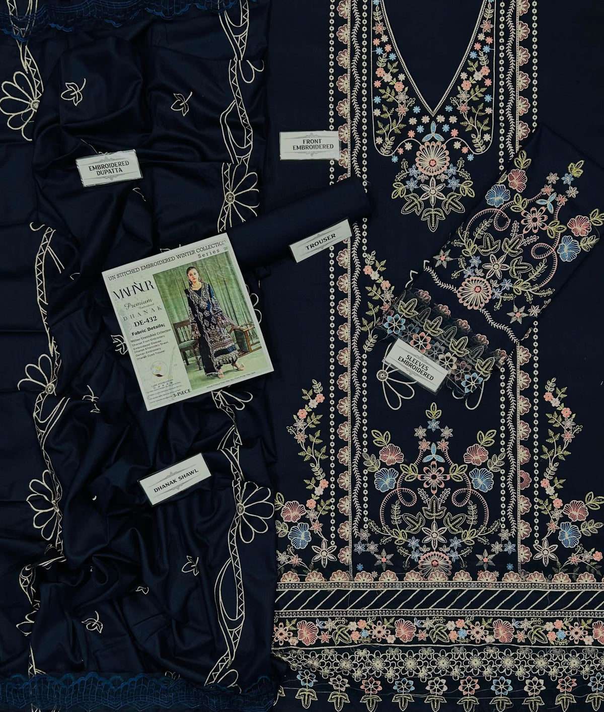 A navy blue embroidered ethnic suit set laid flat, featuring a heavily embellished embroidered neckline panel with floral patterns in pink, gold, and blue, matching embroidered fabric borders, and a folded matching fabric piece. Several small white labels are attached, and a magazine insert is placed on top.