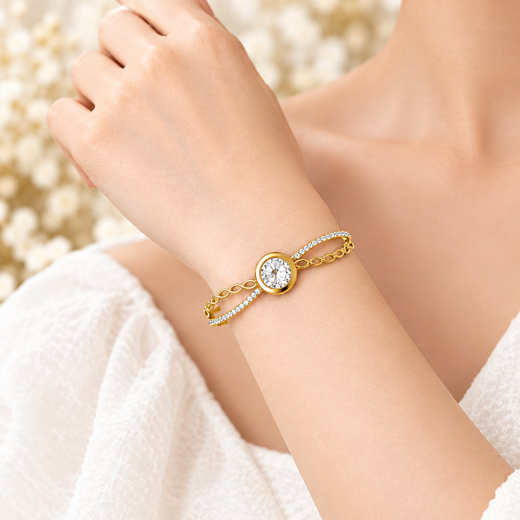A gold bangle bracelet with a central round clear crystal and delicate chain details, displayed on a soft beige fabric background with dried floral accents.