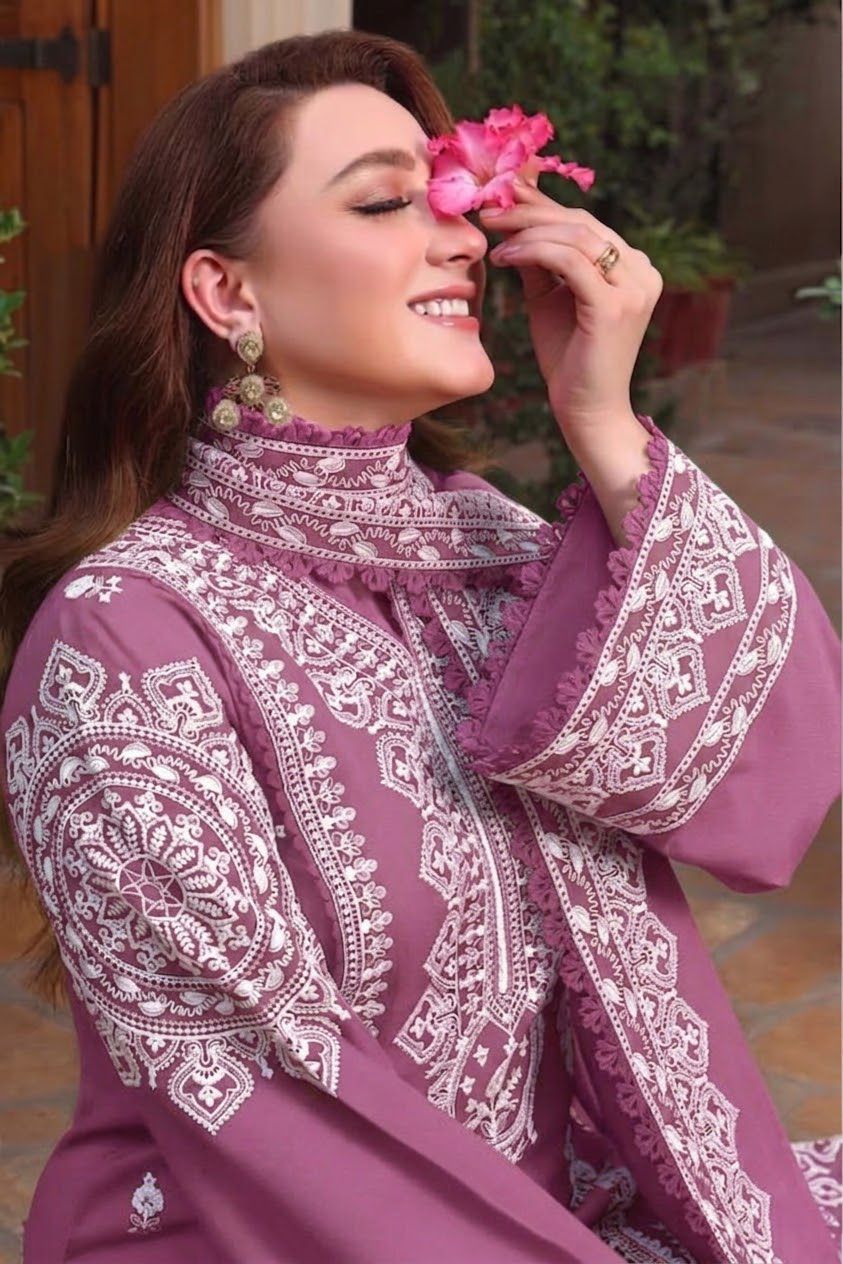 A woman wearing a dusty purple embroidered tunic with white floral patterns, holding a pink flower to her eye in an outdoor setting.