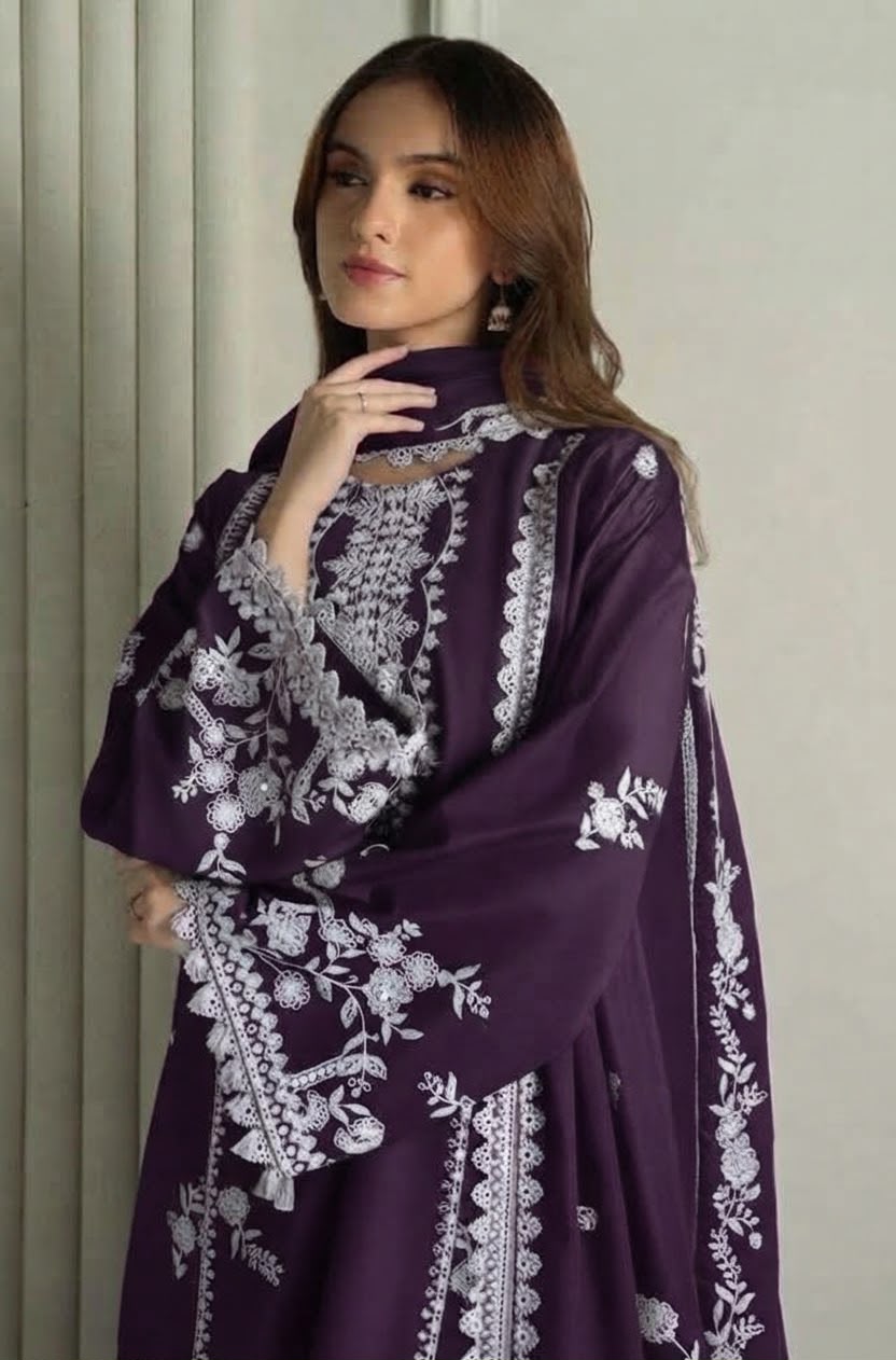A woman wearing a deep purple traditional outfit with intricate white floral embroidery, standing against a neutral beige background.