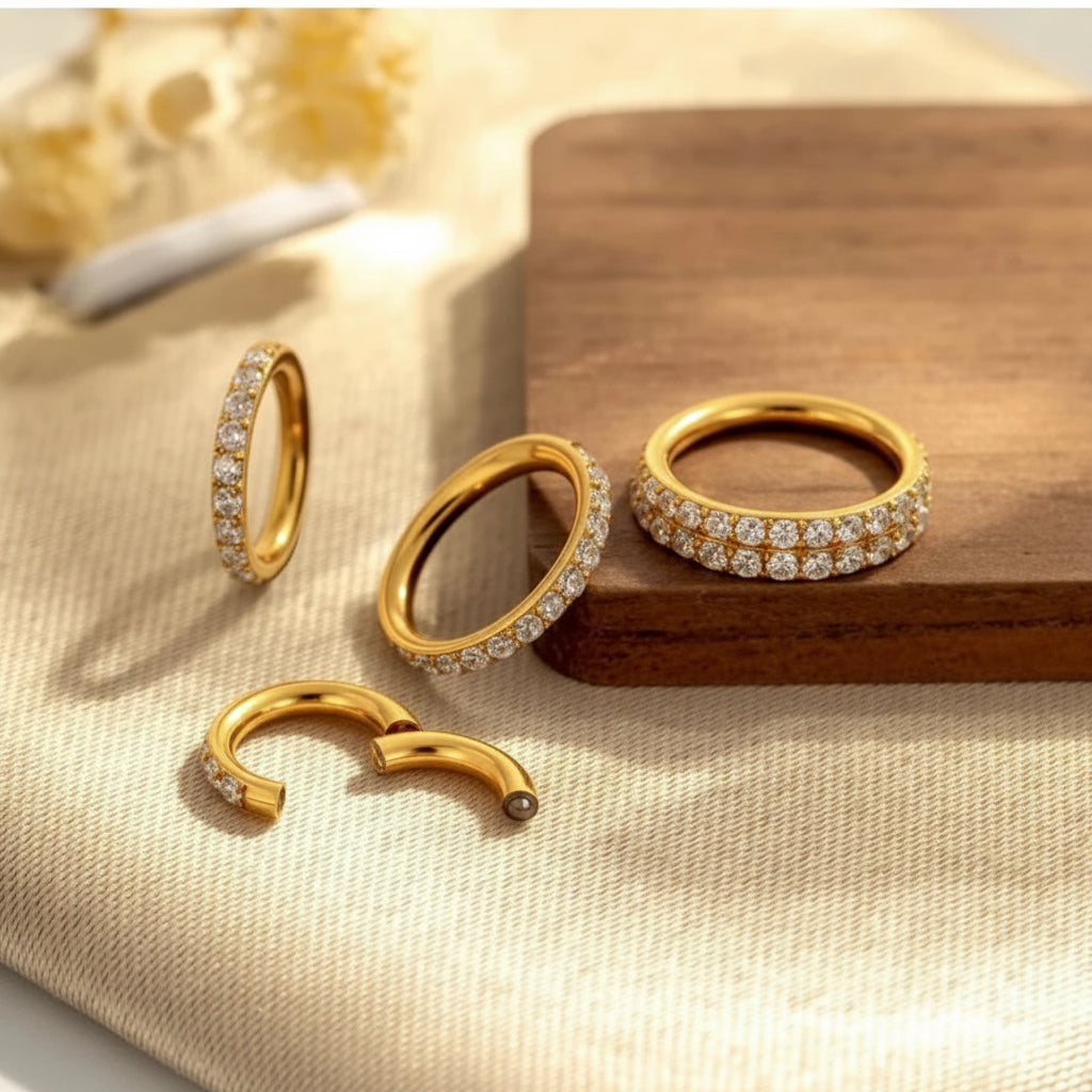 Four gold hoop earrings with clear stones, including two full hoops, one half-hoop, and one open-ended piece, arranged on a beige fabric surface with a wooden block.