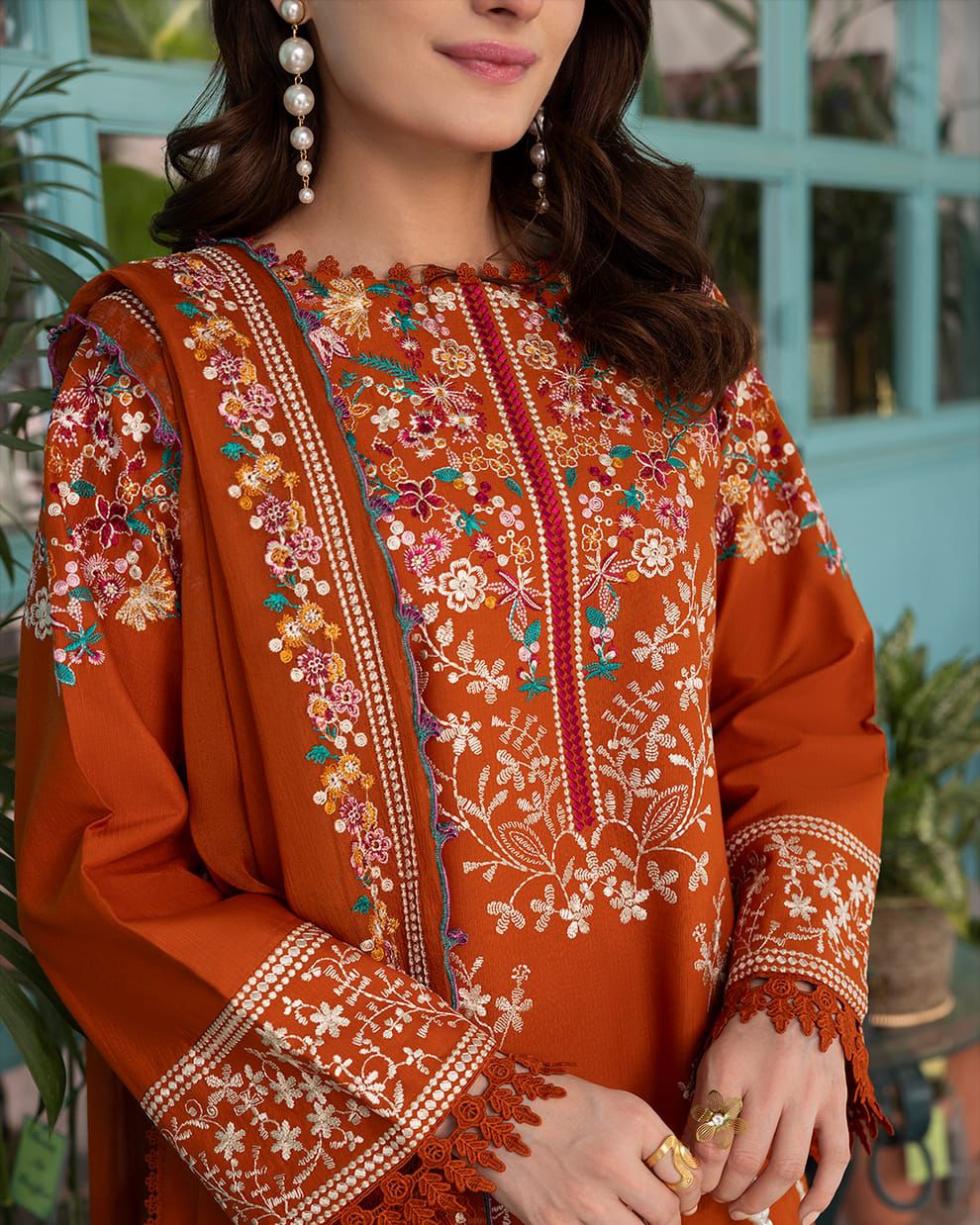 A woman wearing a rust-orange embroidered kurta with floral and geometric patterns, paired with a matching embroidered dupatta, set against a soft-focus background of a turquoise window and green plants.