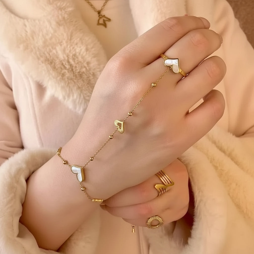 White heart stainless steel hand chain for women
A close-up of a person's hand and wrist wearing delicate gold chain jewelry with heart-shaped charms, including a bracelet and ring, against a soft beige fuzzy background.