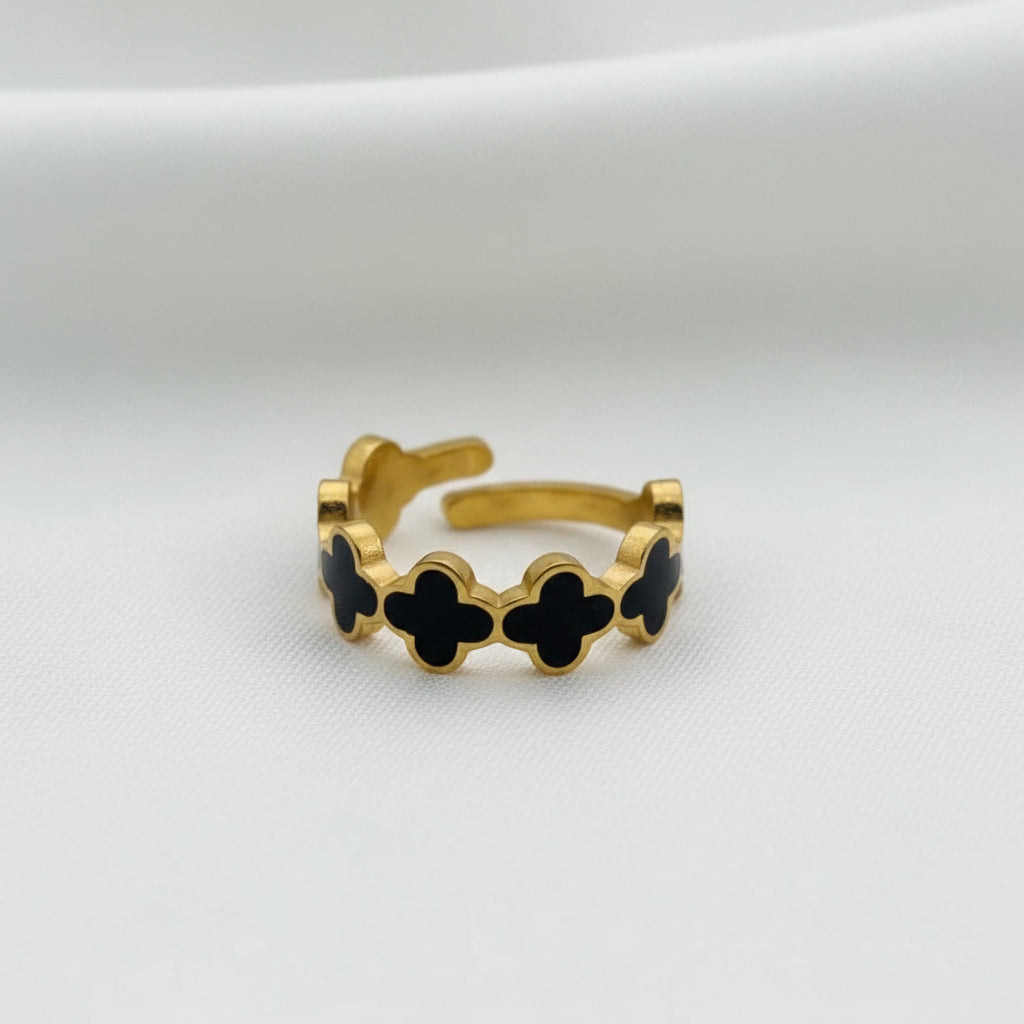 A gold ring featuring four black clover-shaped inlays, displayed on a textured white fabric background.