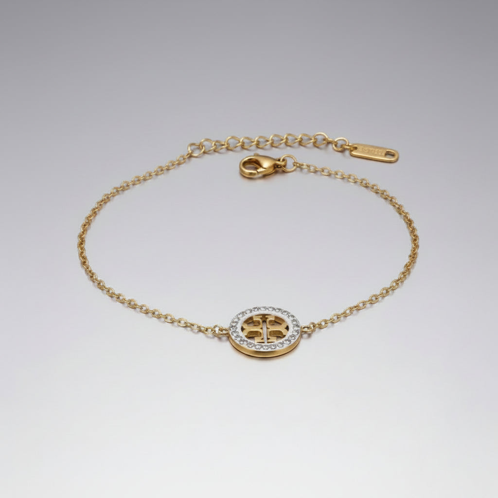 A gold chain bracelet with a circular logo charm, featuring a gold Tory Burch-style monogram center surrounded by a halo of small clear stones, displayed on a white fabric background.