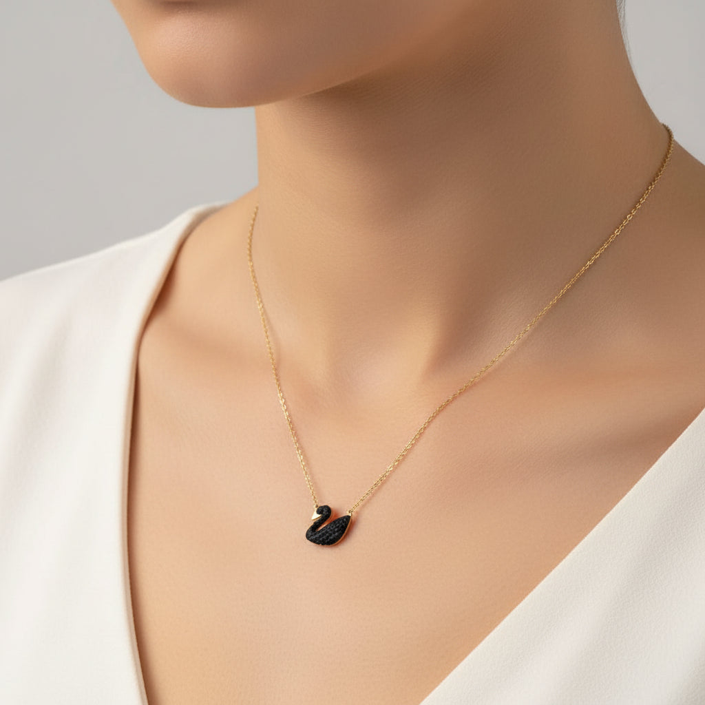 A gold chain necklace with a black swan pendant, resting on a textured white fabric background.