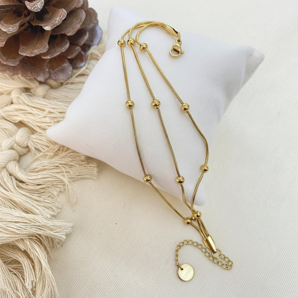 A gold-toned double-strand necklace with small bead accents and a dangling chain with a round tag, displayed on a white fabric cushion with a pinecone and cream fringe prop.