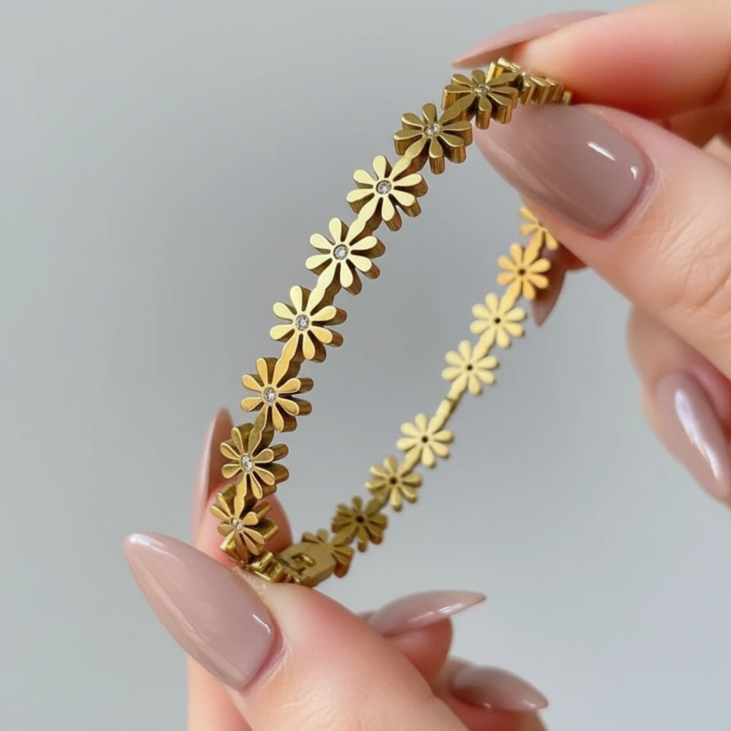 A gold-tone floral bangle bracelet with small flower motifs and tiny clear center stones, held between fingers against a soft neutral background.