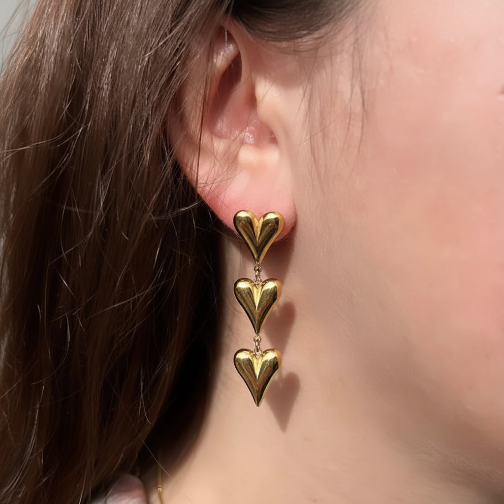 A close-up of a person's ear wearing a gold drop earring with three stacked heart charms.