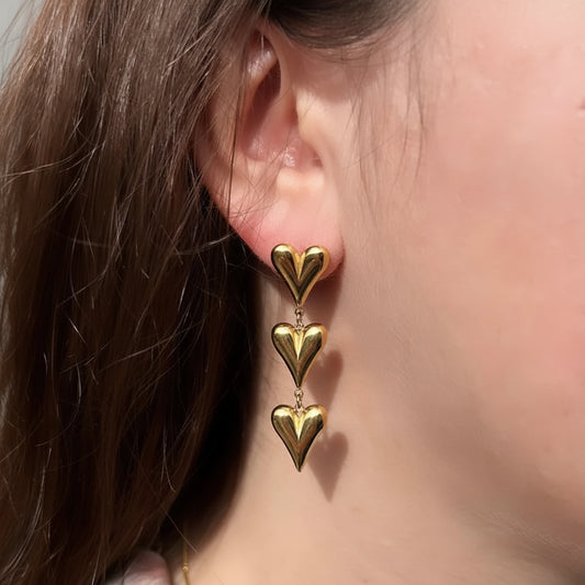 A close-up of a person's ear wearing a gold drop earring with three stacked heart charms.