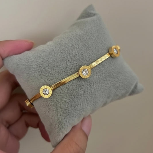 A gold bracelet with three round bezel-set clear stones, displayed on a gray velvet jewelry pillow held by a hand.