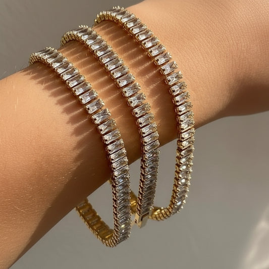 Three gold tennis bracelets with clear baguette-cut rhinestones worn stacked on a person's wrist, shown against a neutral light gray background.
