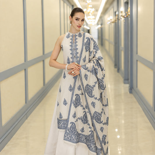 A woman models a traditional white three-piece salwar kameez suit with blue paisley and border prints, standing in a brightly lit hallway.