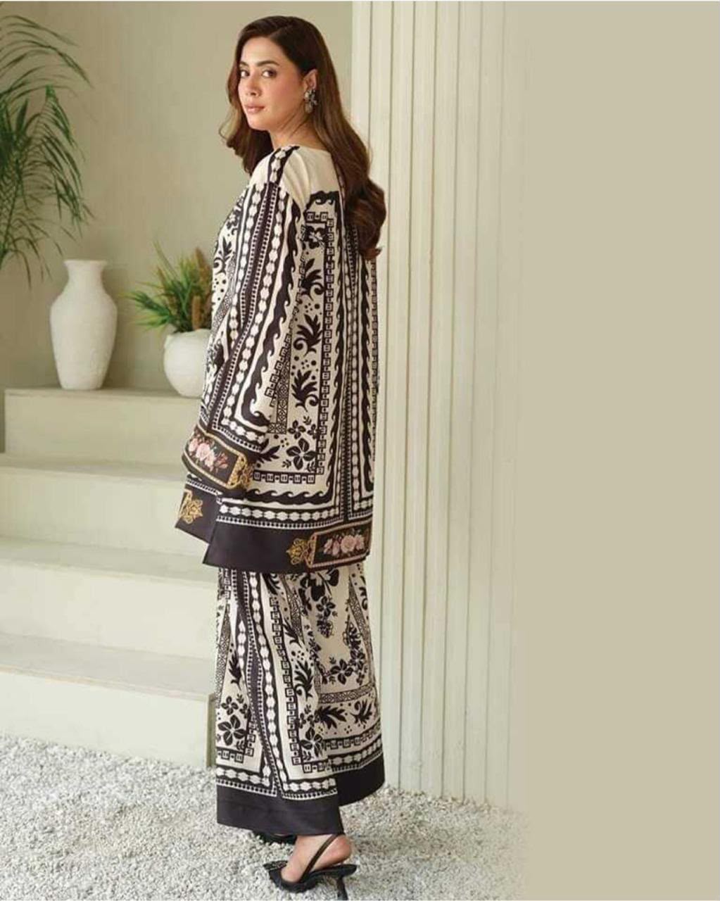 A woman models a three-piece printed ethnic suit in a neutral indoor setting. The suit features a cream base with black floral patterns and brown borders, paired with a matching long jacket and wide-leg trousers.