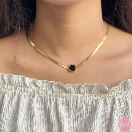 A close-up of a model wearing a gold snake chain necklace with a small round black onyx pendant, paired with a white off-the-shoulder top.