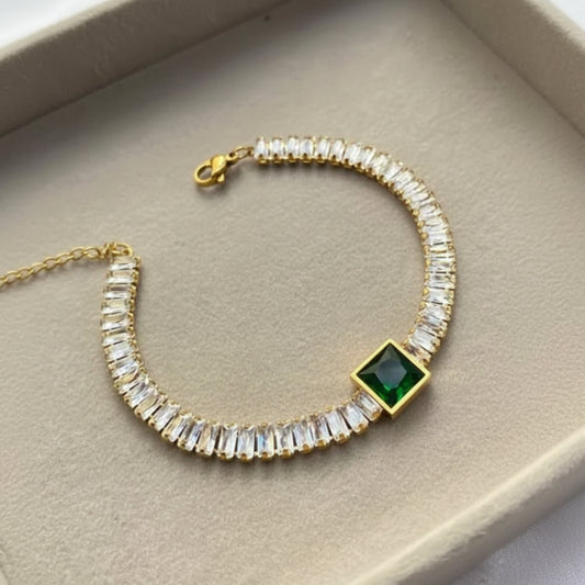 A gold-plated tennis bracelet featuring clear rectangular crystals and a central square green gemstone, displayed inside a beige jewelry box.