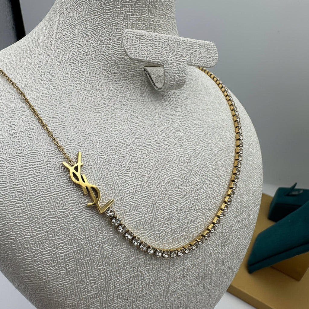 A gold-tone necklace displayed on a textured beige jewelry bust. The necklace features a prominent interlocking YSL logo pendant and a line of clear rhinestones along one side.