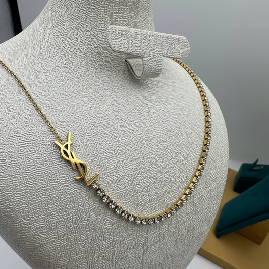 A gold-tone necklace displayed on a textured beige jewelry bust. The necklace features a prominent interlocking YSL logo pendant and a line of clear rhinestones along one side.