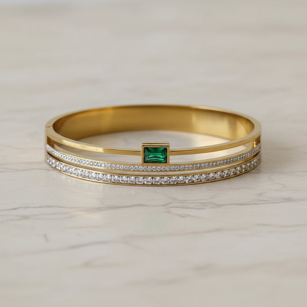 A gold-plated bangle bracelet featuring a central square-cut green emerald-cut gemstone, flanked by two rows of small clear pavé-set stones, displayed on a soft beige fabric background.