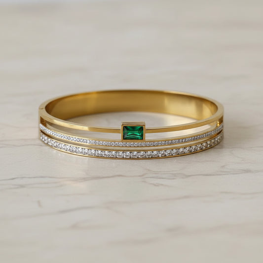 A gold-plated bangle bracelet featuring a central square-cut green emerald-cut gemstone, flanked by two rows of small clear pavé-set stones, displayed on a soft beige fabric background.