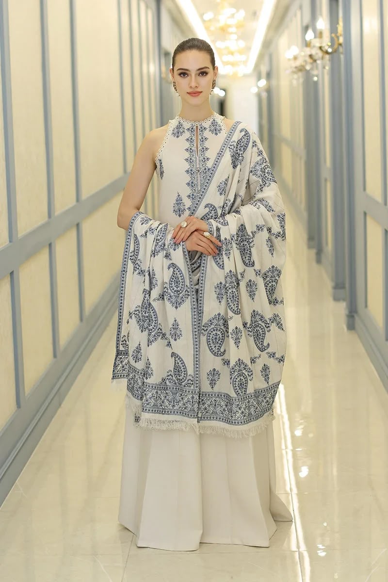 A woman models a traditional white three-piece salwar kameez suit with intricate blue floral embroidery, standing in a brightly lit hallway with cream walls and blue trim.