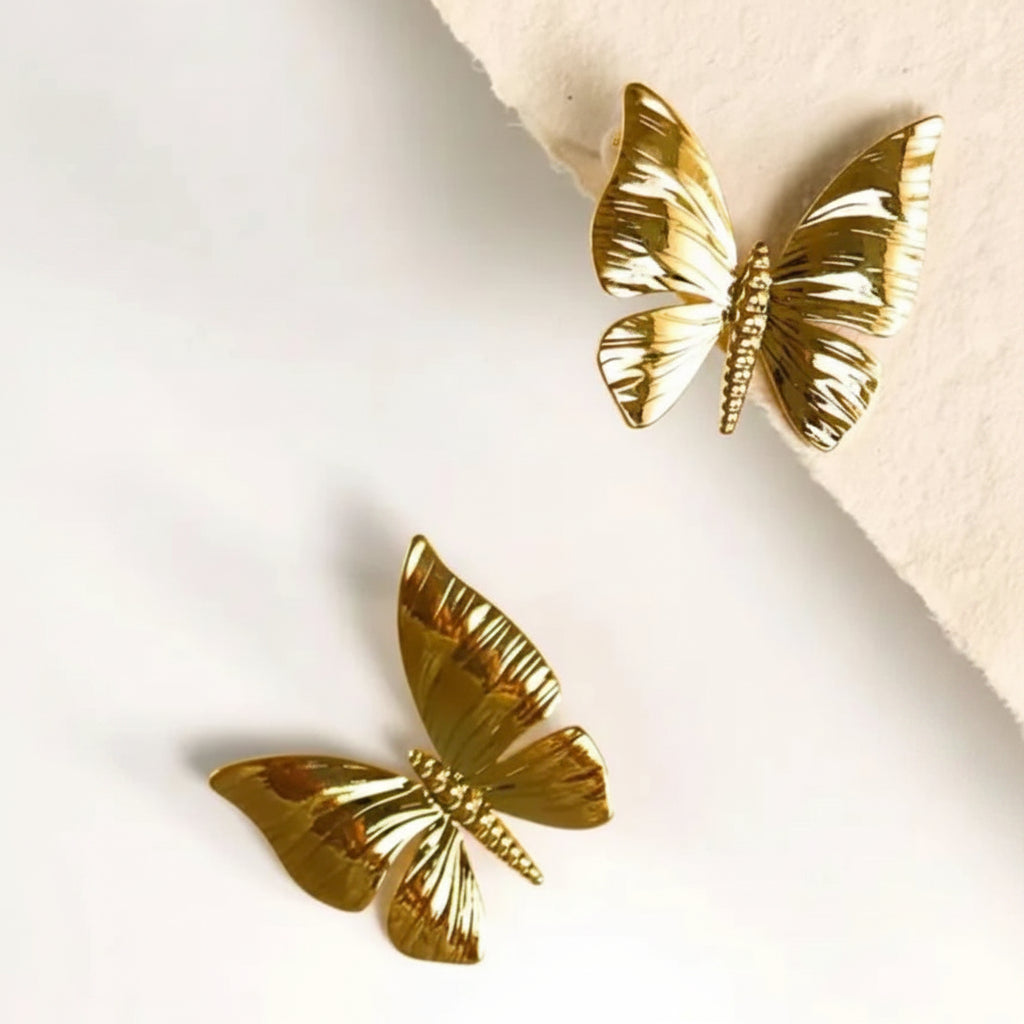 Two gold butterfly-shaped earrings with textured wings and beaded bodies, displayed on a white surface with a beige paper corner.