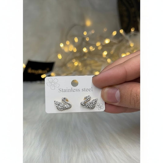 A hand holding a white card displaying a pair of silver-toned swan-shaped stud earrings covered in clear rhinestones, with a blurred warm light background.