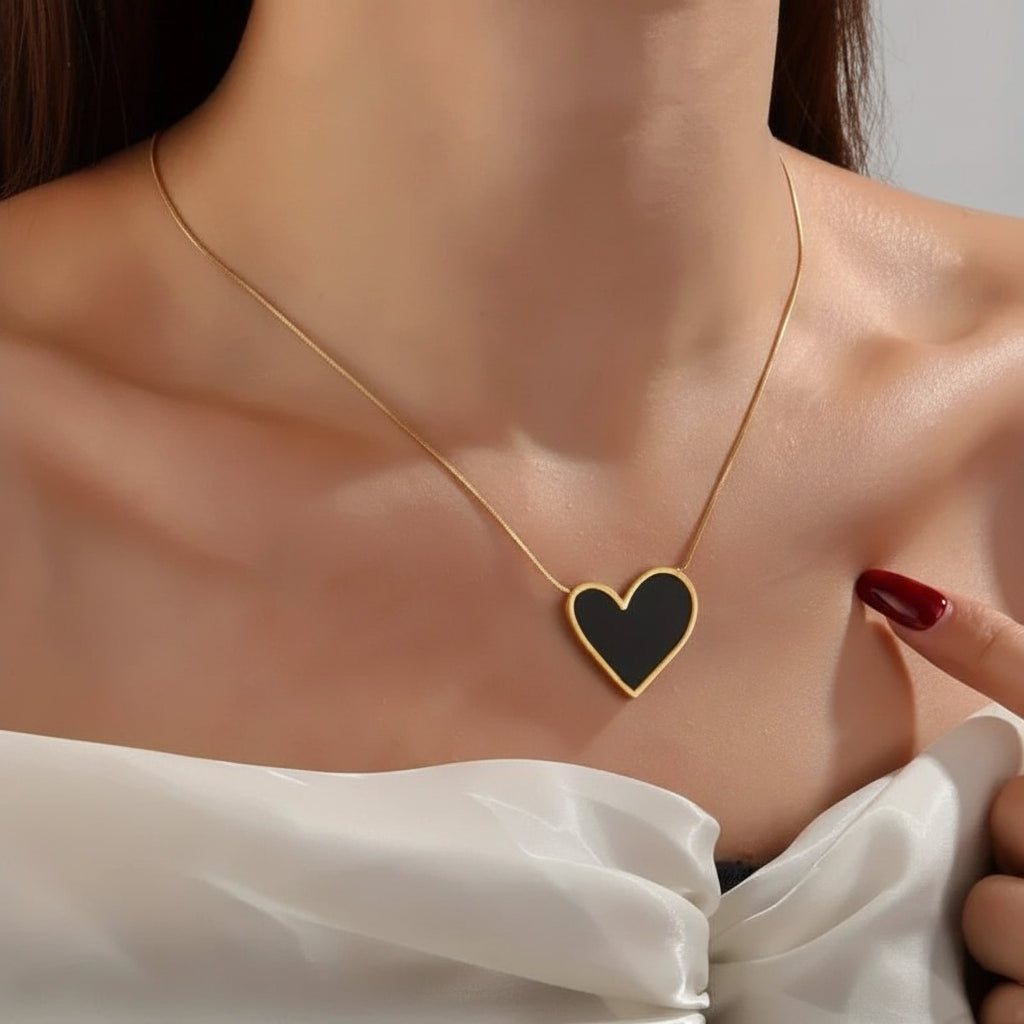 A close-up of a model wearing a delicate gold chain necklace with a heart-shaped pendant. The pendant features a matte black center framed by a polished gold border.